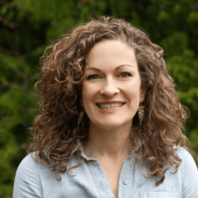 A person with curly hair and a light blue shirt, standing in front of green foliage, smiles at the camera, embodying the fresh approach of our senior living advisory service.